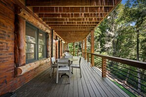 A wooden deck with a table and chairs, surrounded by a wooden railing, overlooking a forested area. - Four Seasons Mountain Escape - Riverfront | Pet Friendly | Hot Tub (Blue Ridge)