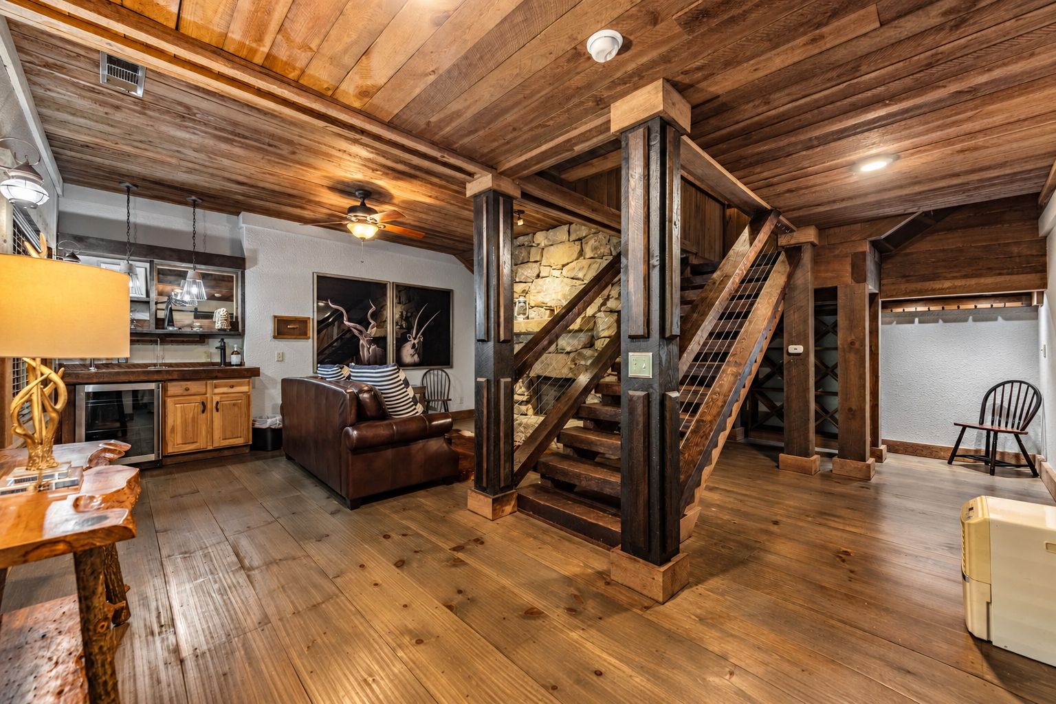 A spacious living area with a wooden staircase, a kitchen in the background, and a stone wall.