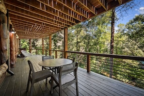 A wooden deck with a table and chairs, surrounded by a railing, overlooking a forested area. - Four Seasons Mountain Escape - Riverfront | Pet Friendly | Hot Tub (Blue Ridge)