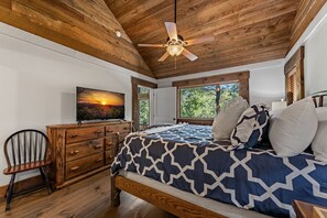A bedroom with a wooden ceiling, a bed with patterned bedding, a television on a wooden dresser, and a window with a view of trees. - Four Seasons Mountain Escape - Riverfront | Pet Friendly | Hot Tub (Blue Ridge)