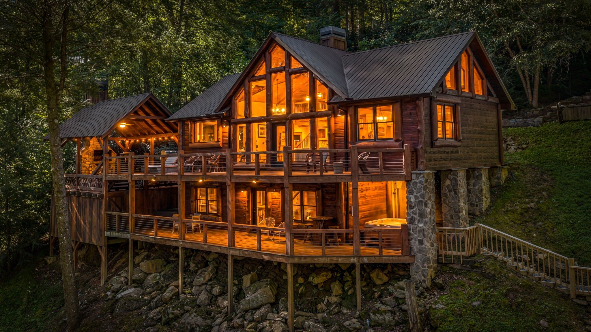 A multi-level wooden cabin with a stone foundation, featuring a balcony and a hot tub.