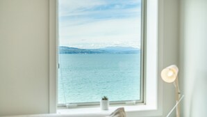 Ein Zimmer mit großem Fenster, das auf ein Gewässer und ferne Berge blickt.
