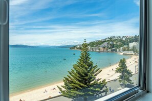A beach view from a window, featuring a clear blue sea, sandy beach, and a row of buildings in the background. - Oriental Bay Waterfront- Entire 2 Level apartment (Wellington)