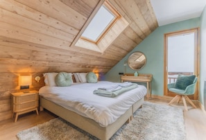 Ein Schlafzimmer mit schrägen Wänden aus Holz, einem Bett mit Kissen, einem Nachttisch mit Lampe, einem Spiegel, einem Sessel und Blick durch ein Fenster.