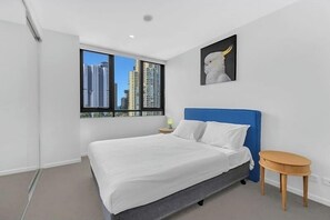 A modern bedroom with a large bed, a blue headboard, a small wooden table, a nightstand, a city view through a window, and a wall-mounted artwork of a white bird. - Modern 2BR Walking Distance to Beach with Parking (Surfers Paradise)
