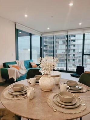 A modern dining area with a round table set for four, surrounded by a large window with a city view. - Stylish 1BR | Pool, Gym and Parking by Olympic Park (Lidcombe)