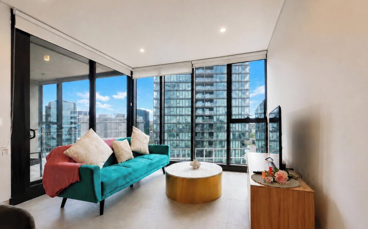 Un salon moderne avec un canapé de couleur turquoise, une table basse ronde et une vue sur les gratte-ciel, visible à travers de grandes fenêtres.