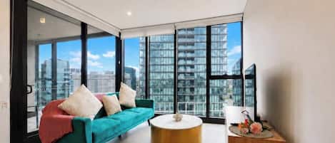 A modern living room with a teal sofa, a round coffee table, and a view of high-rise buildings through large windows.
