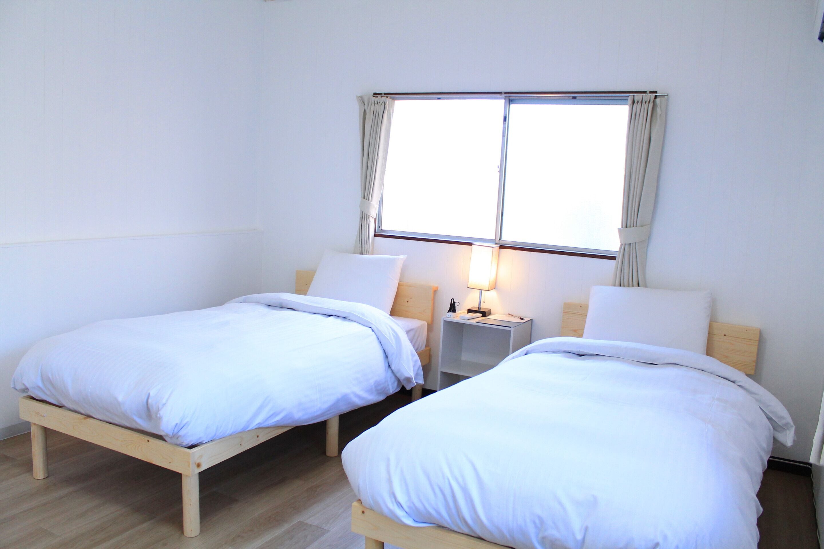 Dos camas individuales con estructuras de madera y ropa de cama blanca, una mesita de noche con lámpara y una ventana con cortinas.