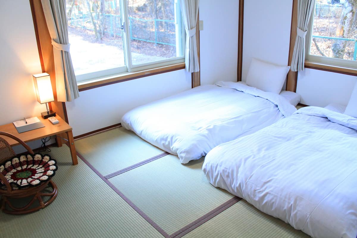 A room with two beds, a desk with a lamp, and a view of the outdoors through the window.