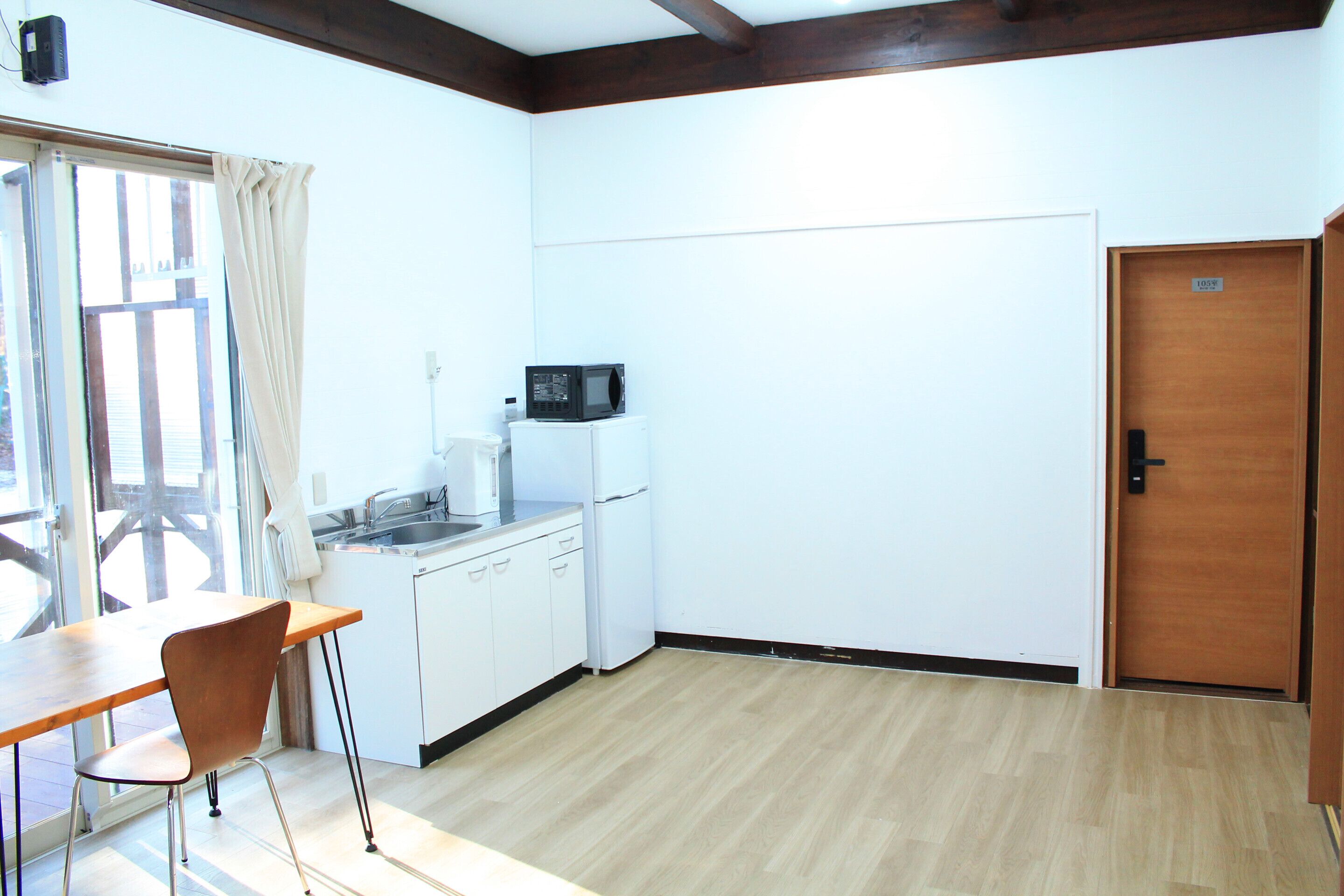 A small kitchen with a microwave, sink, and white cabinets. There is a wooden table and a chair, and a door with a number sign.