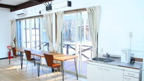 Ein Zimmer mit einem Holzespeisetisch, Stühlen, einer Küchenzeile mit Spüle und Schränken sowie großen Fenstern mit Vorhängen.