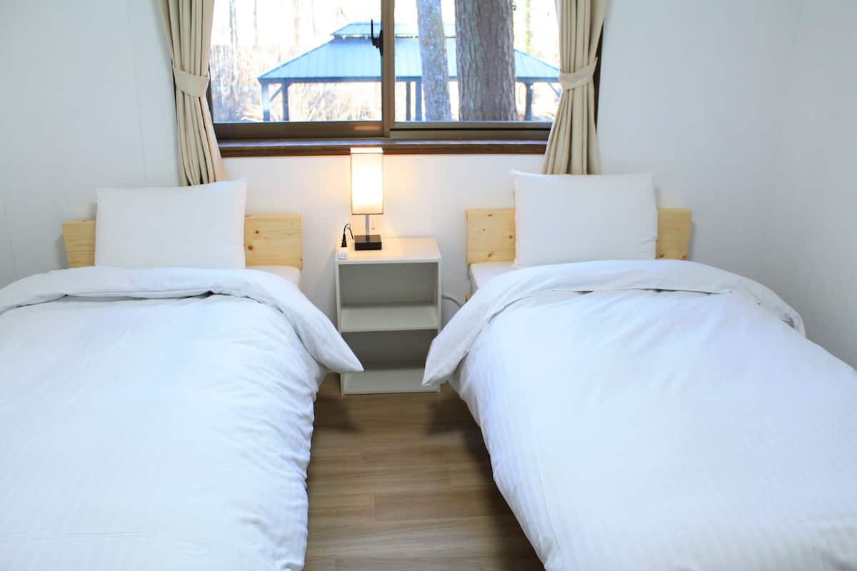 A twin room with two beds, a nightstand with a lamp, a window with curtains, and a view of the outdoors.