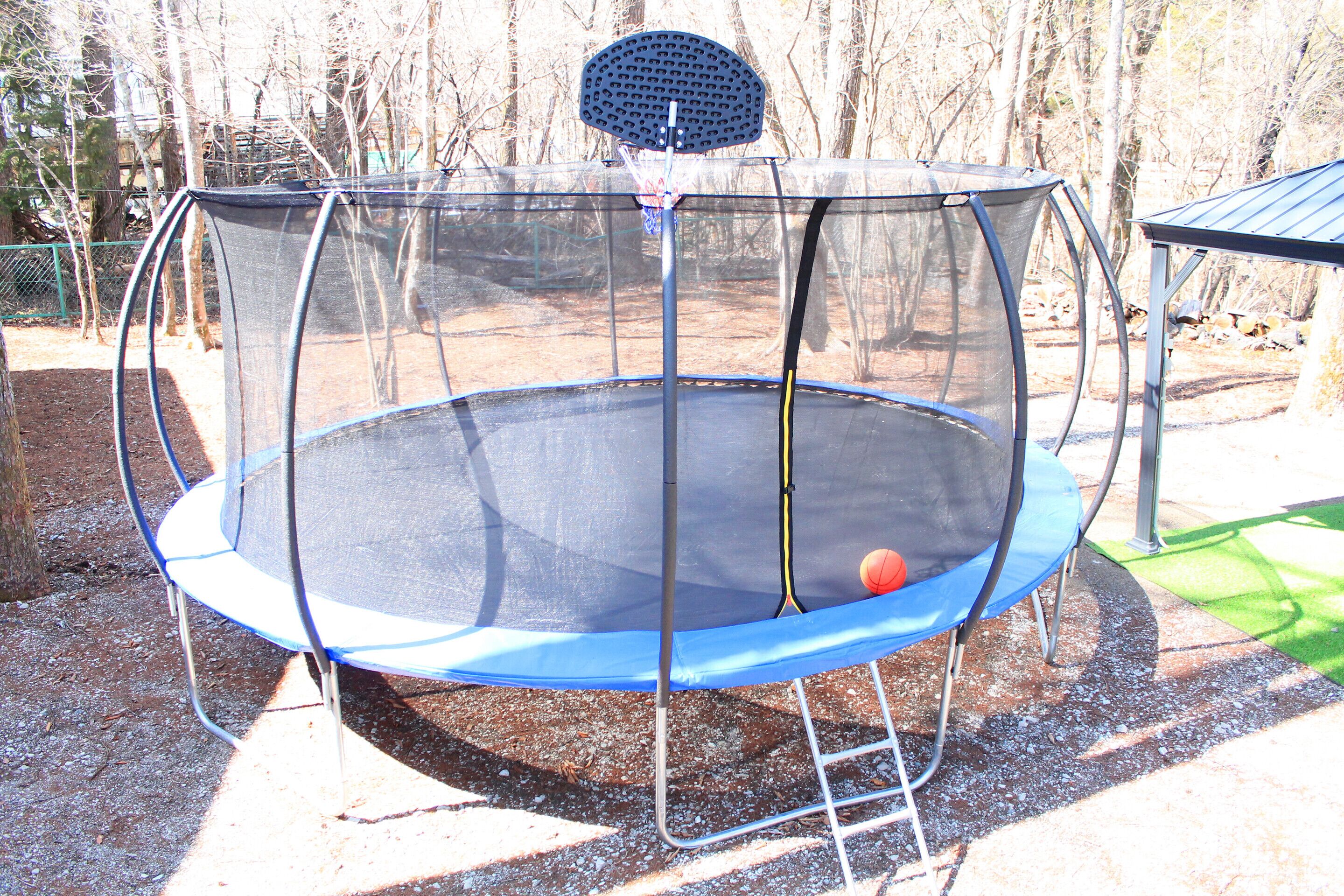 A trampoline with a safety net enclosure, a basketball hoop, and a shaded area.