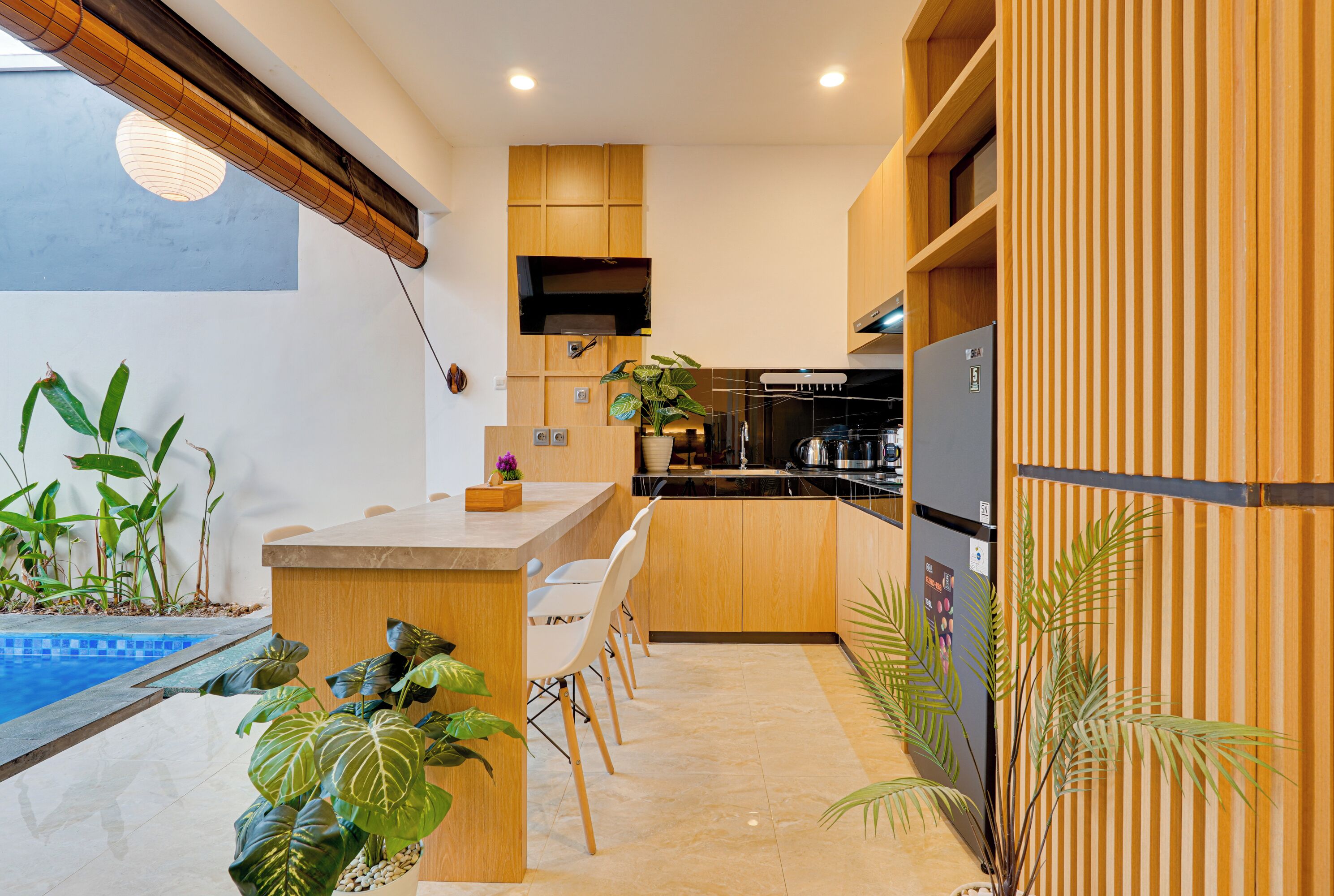 Cozinha moderna com armários de madeira, ilha integrada com assentos, geladeira e pequena área de piscina interna com plantas.