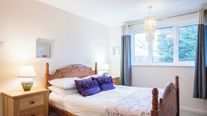 Ein ordentlich bezogenes Bett mit Holzkopfteil, zwei Nachttische mit Lampen, ein Fenster mit Vorhängen und ein Kronleuchter.