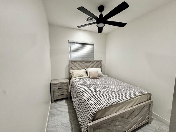 Un dormitorio con una cama, una mesita de noche, un ventilador de techo y una ventana con persianas.