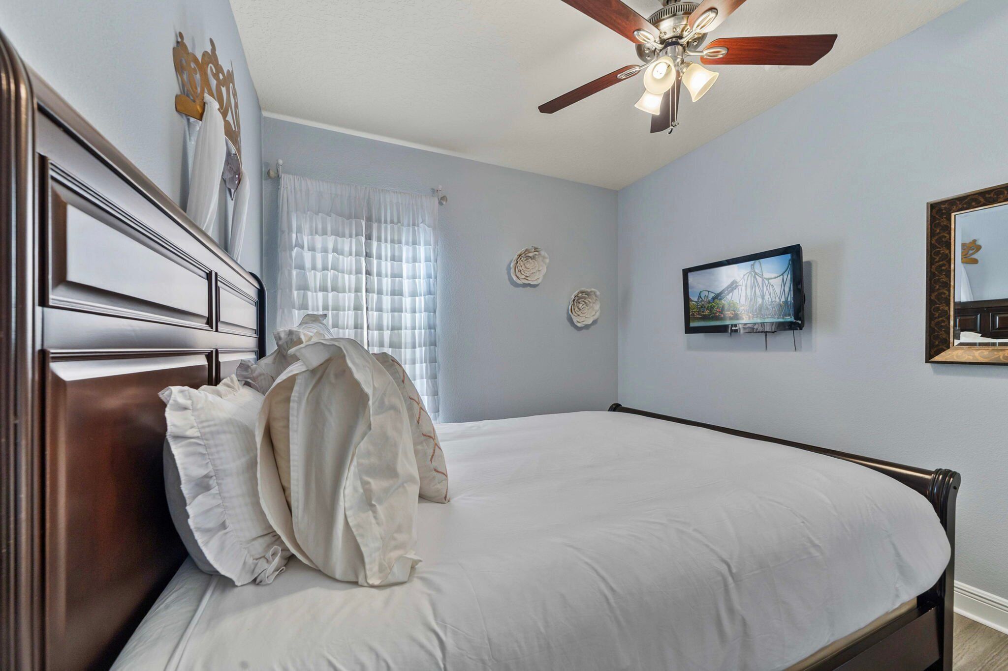 A neatly made bed with white linens, a ceiling fan, a television on the wall, and decorative wall art.