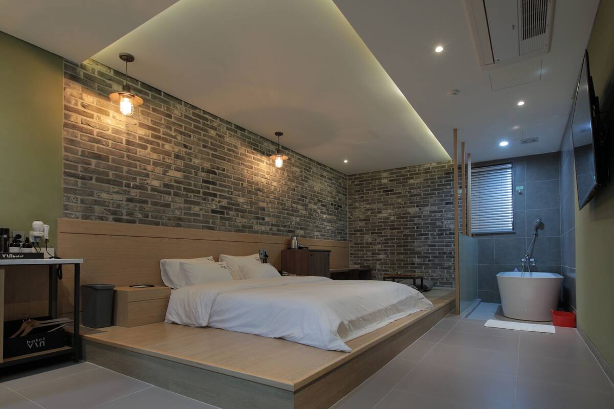 Une chambre d’hôtel moderne avec un mur en briques, un grand lit, une table de chevet en bois, une télévision et une salle de bain avec une baignoire.