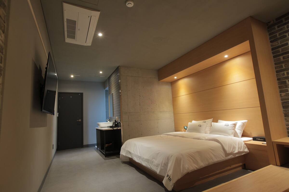 Une chambre d’hôtel moderne avec un grand lit, une table de chevet, un téléviseur fixé au mur et une salle de bain équipée d’un lavabo et d’un miroir.