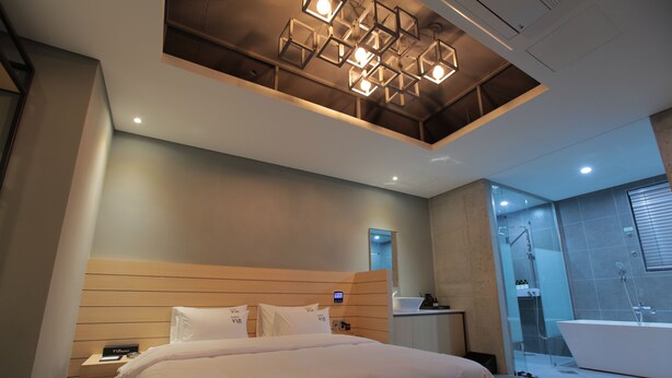A modern hotel room with a large bed, a wooden headboard, a built-in desk, a vanity with a mirror, and a separate shower area. - Hotel Vin (Changwon)