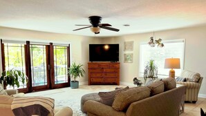 Una sala de estar con sofá, televisor, ventilador de techo y ventana con persianas.