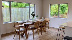 Un espace repas avec une table en bois et des chaises en osier, de grandes fenêtres donnant sur un paysage verdoyant et une cuisine moderne.