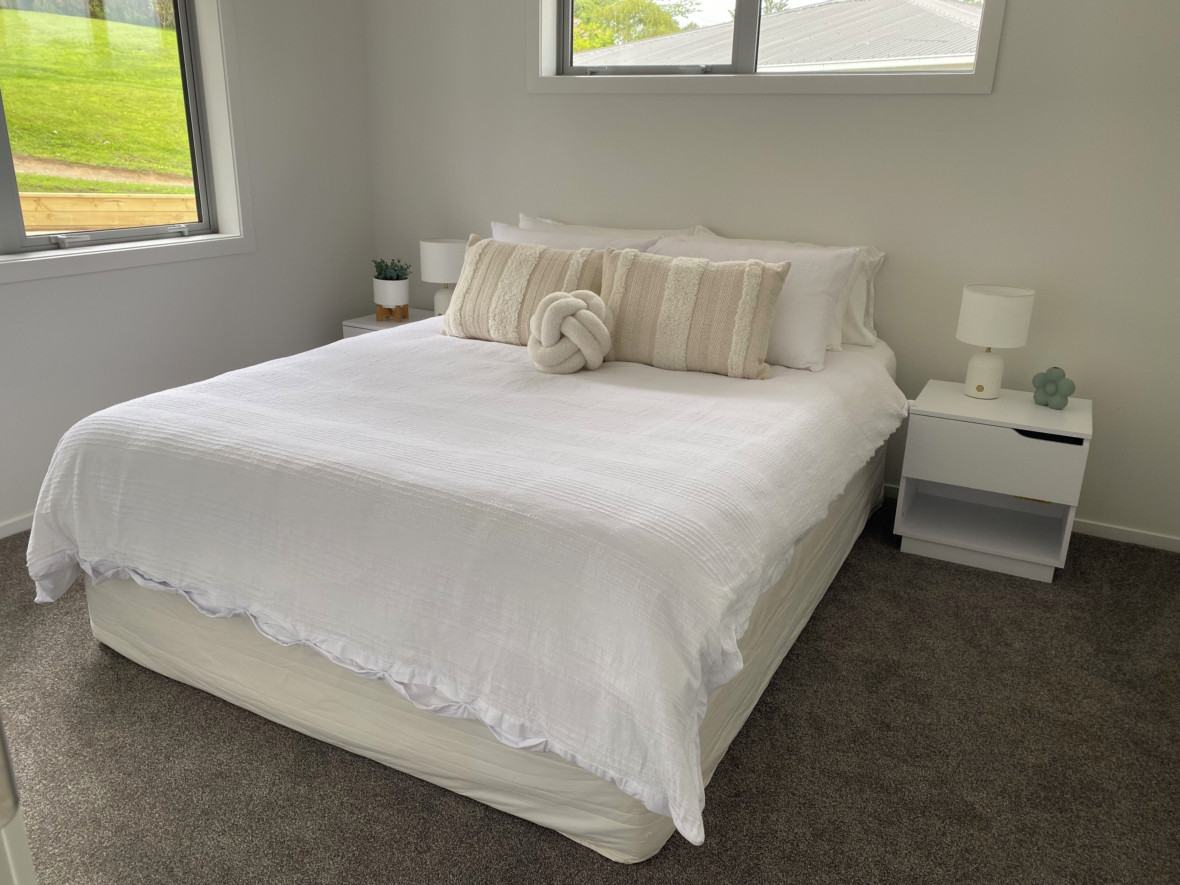Uma cama bem arrumada com edredom branco e almofadas, uma mesa de cabeceira com abajur e vista para um gramado verde pela janela.