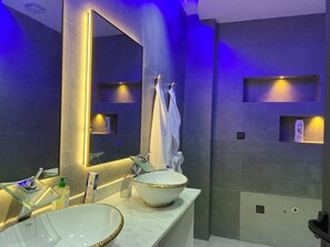 Un baño moderno con un espejo grande, un lavamanos blanco con detalle dorado y una toalla blanca colgada en un perchero.