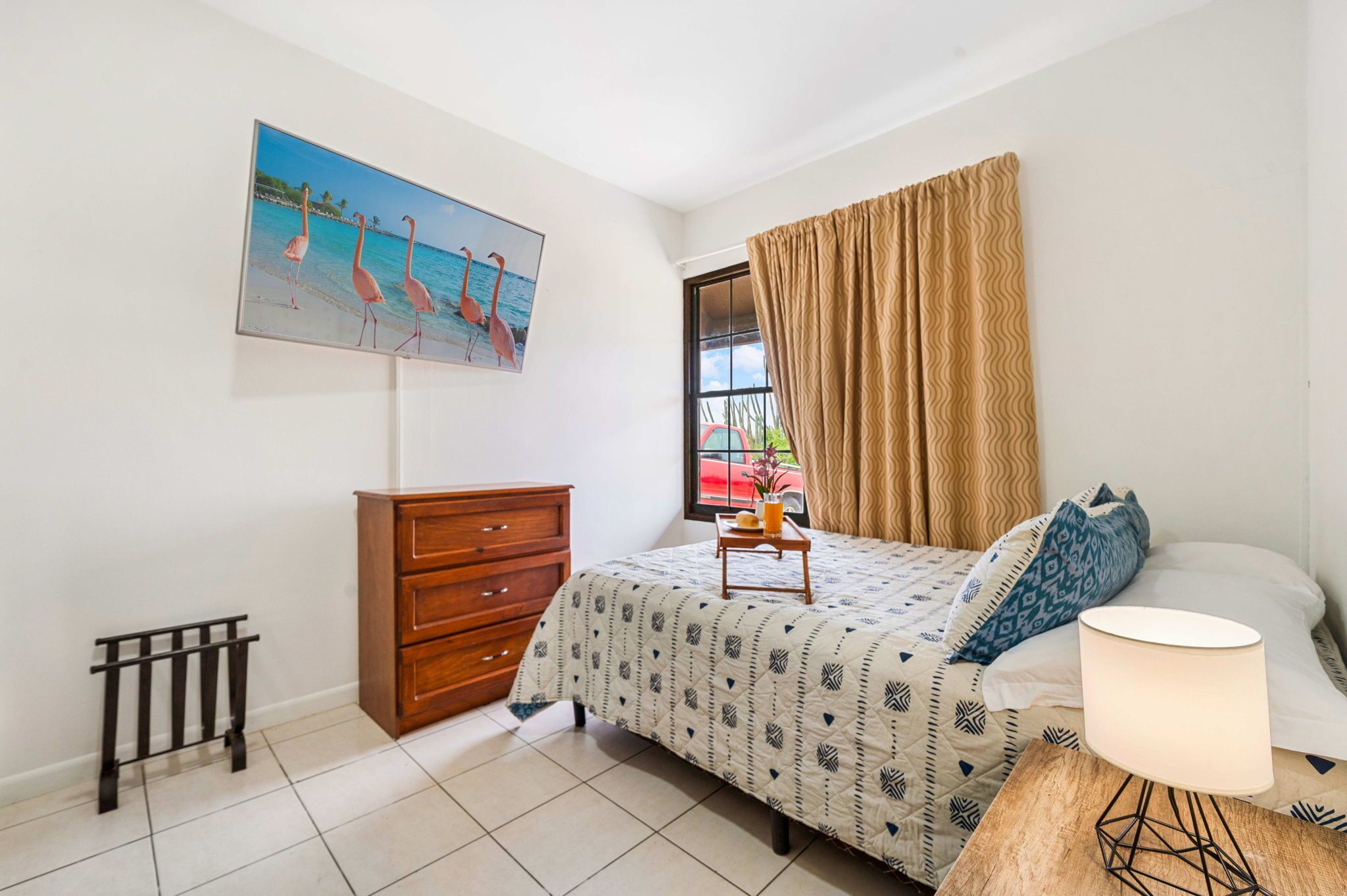 Une chambre à coucher avec un lit, une commode, un radiateur, un tableau représentant des flamants roses et une fenêtre avec des rideaux.