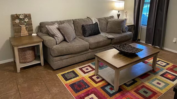 A living room with a grey sofa, a wooden coffee table, a colorful rug, a wall-mounted welcome sign, and a lamp. - Cozy Pet-Friendly 3BR Duplex with Fast WiFi only Minutes to Casino (Fort Smith)