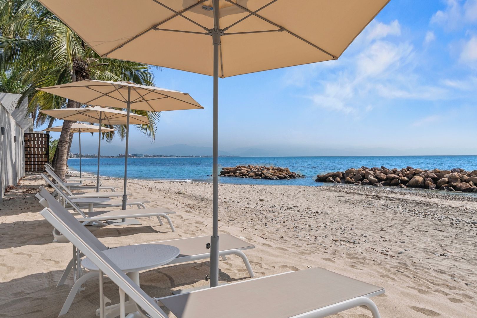 Ein Sitzbereich am Strand mit Sonnenschirmen und Liegestühlen, eine felsige Küstenlinie und ein freier Blick auf das Meer.