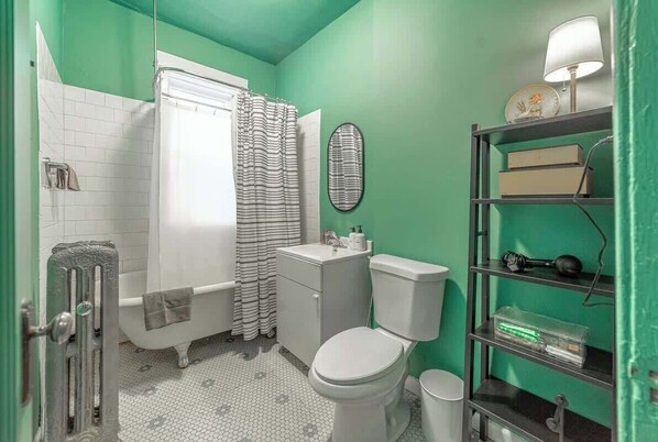 A bathroom with a white bathtub, a toilet, and a shower curtain. There is a shelf with various items and a mirror on the wall. - The Codfather at Wrigley (Chicago)