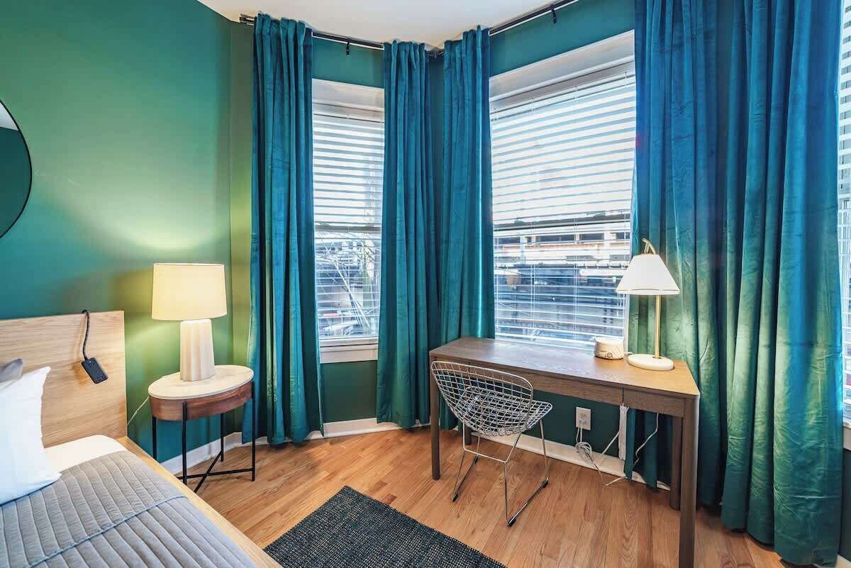 Une chambre avec un lit, un bureau, deux lampes, une chaise et de grandes fenêtres avec des rideaux.