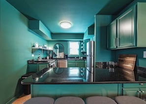 A modern kitchen with teal cabinetry, a central island, and a stainless steel refrigerator. - The Ivy Flat | Designer 4BR Steps to Wrigley Field (Chicago)