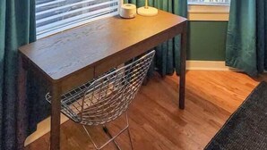 A corner room with a wooden desk, a chair, a lamp, and a window with blinds. - The Ivy Flat | Designer 4BR Steps to Wrigley Field (Chicago)