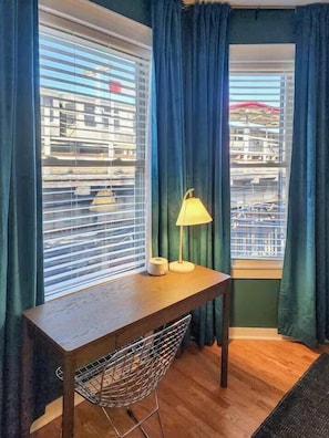 A corner room with a wooden desk, a chair, a lamp, and a window with blinds. - The Ivy Flat | Designer 4BR Steps to Wrigley Field (Chicago)