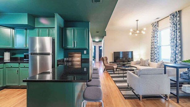A modern kitchen with teal cabinets and a central island, leading to a living area with a sofa and TV. - The Ivy Flat | Designer 4BR Steps to Wrigley Field (Chicago)