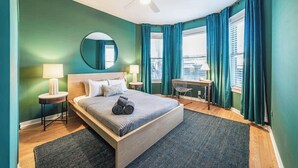 A modern bedroom with a bed, two bedside tables, a desk, a chair, a ceiling fan, and a large window with curtains. - The Ivy Flat | Designer 4BR Steps to Wrigley Field (Chicago)