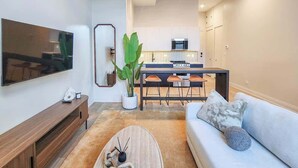 Un salon moderne avec un téléviseur à écran plat, une table basse en bois, un canapé bleu clair et un coin repas avec une cuisine en arrière-plan.