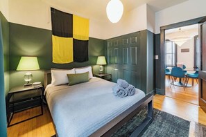 Ein Schlafzimmer mit einem Bett, zwei Nachttischen, einer Lampe, einer Flagge und Blick in einen weiteren Raum mit Tisch und Stühlen.