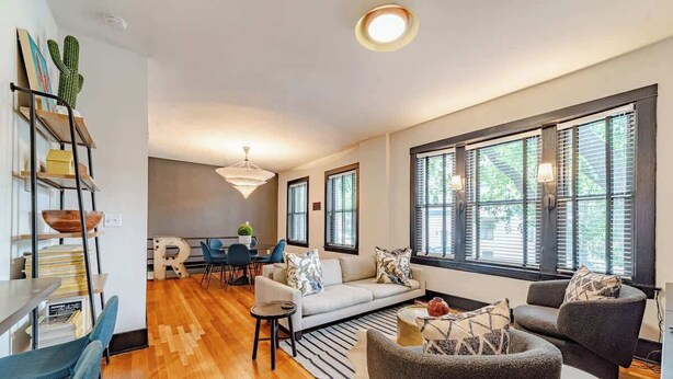 A modern living room with a sofa, armchairs, a coffee table, and a dining area with a table and chairs. There are large windows with blinds, a bookshelf, and a chandelier. - Feelin’ Nauti Near Wrigley (Chicago)