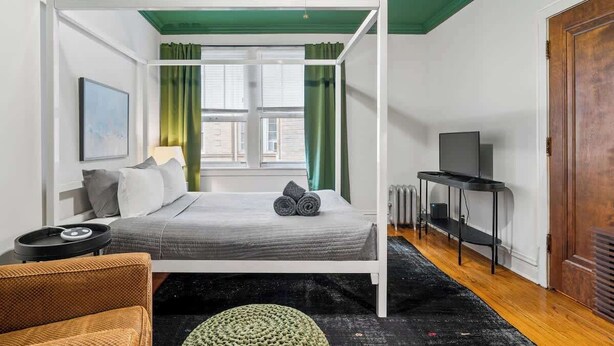 Una camera da letto con un letto, una ventola a soffitto, una finestra con tende, un televisore su un mobile e una porta.