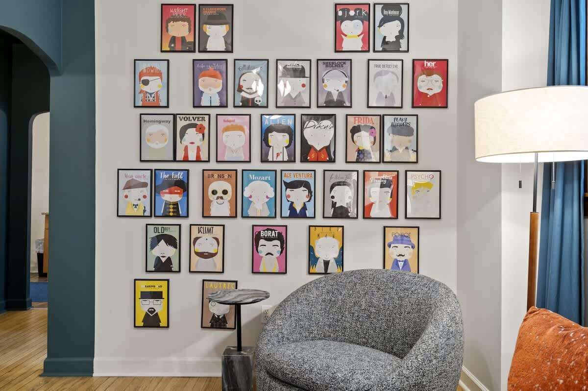 Une pièce dont un mur est décoré d’affiches encadrées, avec un fauteuil gris, une petite table d’appoint et une lampe.