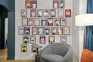 A room with a wall decorated with framed posters, a grey armchair, a small side table, and a lamp. - A Hand-some Place Near Wrigley (Chicago)