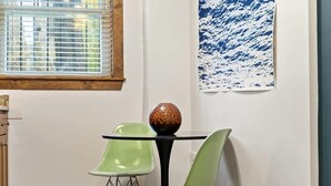 Et spiseområde med et rundt bord, to grønne stole, en dekorativ vase, et vindue med rullegardiner og et abstrakt, blåt maleri på væggen.