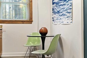 A dining area with a round table, two green chairs, a decorative vase, a window with blinds, and a blue abstract painting on the wall. - A Hand-some Place Near Wrigley (Chicago)