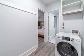 Ein Waschraum mit einer Frontlader-Waschmaschine, einem Wäscheständer und einer Tür zu einem Schlafzimmer mit Bett und Nachttisch.