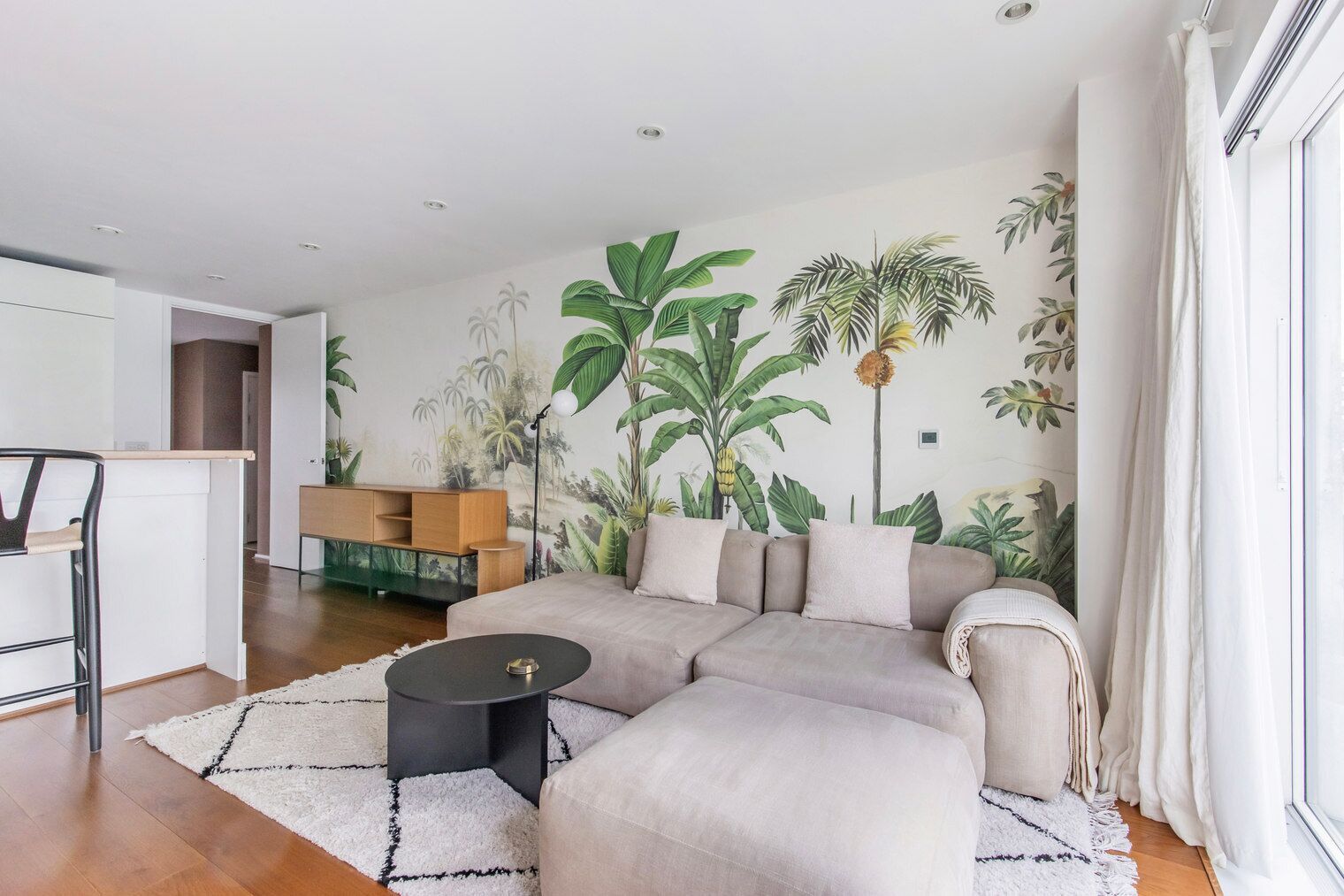 A modern living room with a sectional sofa, a coffee table, and a mural of tropical plants on the wall.