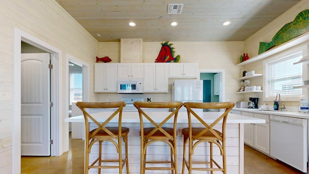 A kitchen with white cabinets, a microwave, a refrigerator, and three wooden bar stools. - GV634, Shared Pool, Pet Friendly, Charcoal Grill, Close to Beach (Port Aransas)